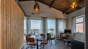 A modern living room with a round dining table, comfortable seating, a flat-screen TV, and large windows offering a city view.