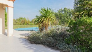 A pool area with a clear blue sky, surrounded by lush greenery and a building with a tiled roof. - Panoramic Poolside Villa (La Croix-Blanche)