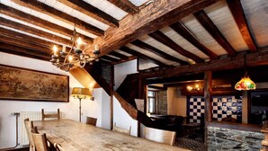 A rustic dining area with a wooden table and chairs, exposed wooden beams, a chandelier, and a stone wall in the background.
