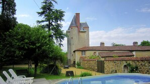 Uma área de piscina com espreguiçadeiras, um edifício de pedra com torre e paisagem gramada.