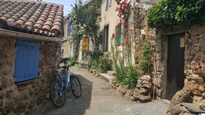 Une ruelle étroite, avec un vélo bleu garé sur le côté gauche, des bâtiments en pierre à l’architecture traditionnelle et un petit jardin agrémenté de fleurs rouges.