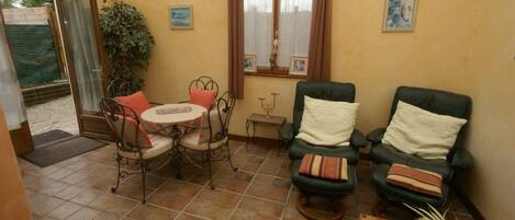 A sunroom with a tiled floor, a table with chairs, two armchairs, a small table with a candle, and a potted plant.