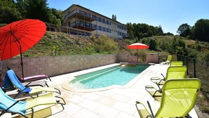 Une piscine entourée de chaises longues et de parasols, le tout bordé d’un mur de pierre et avec un bâtiment visible au loin.