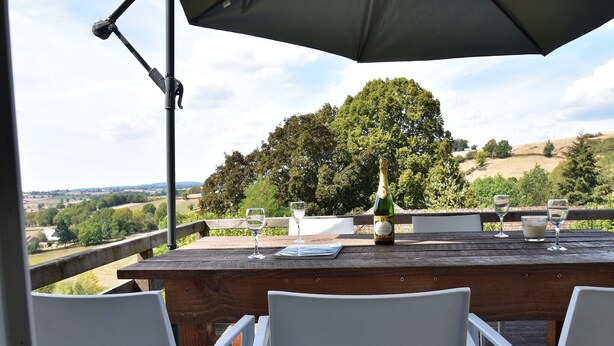 Une table en bois, avec une bouteille et des verres, entourée de chaises blanches, donnant sur un paysage pittoresque.