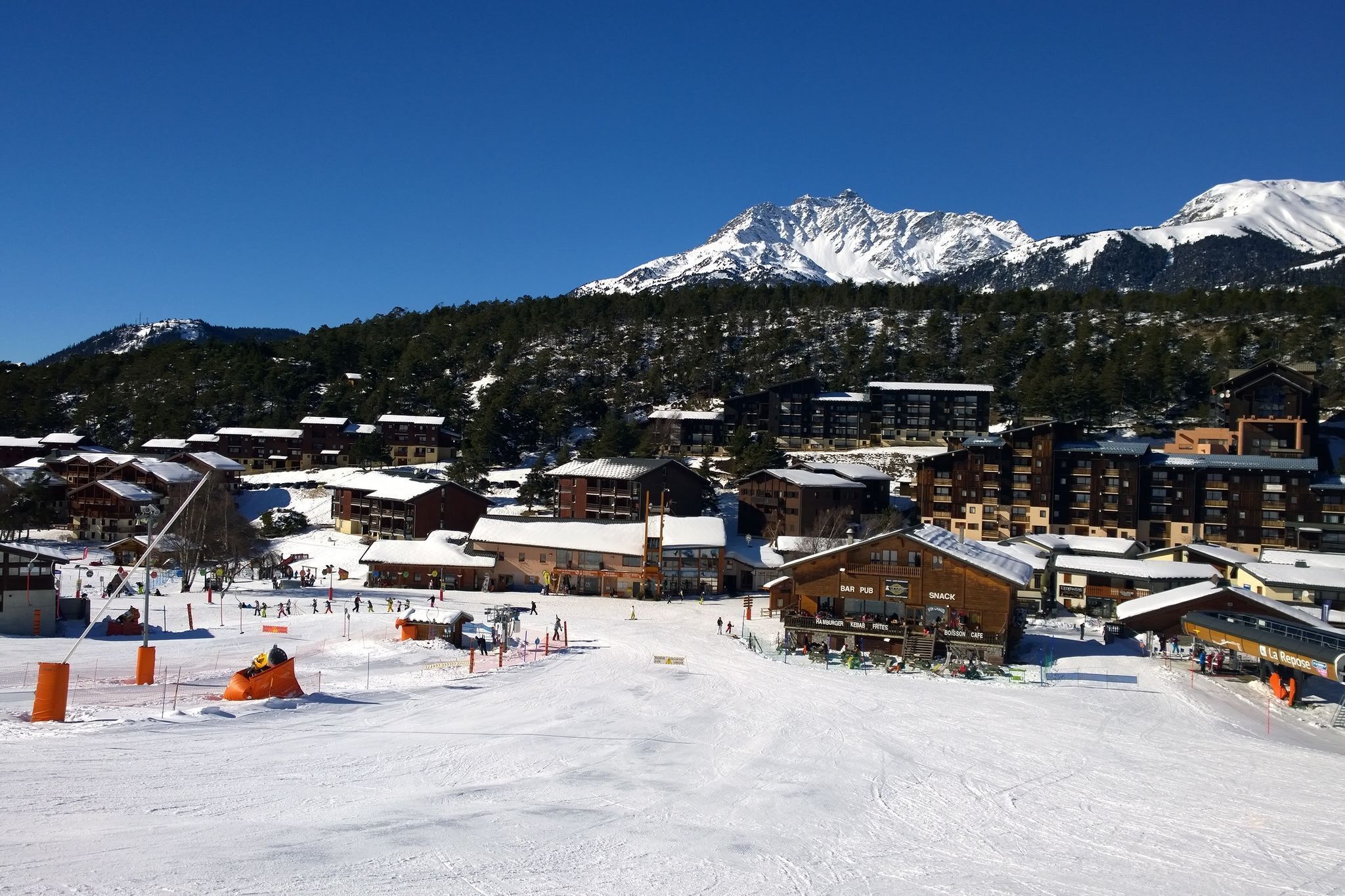 Een bergresort vol sneeuw, met houten gebouwen, skipistes en een heldere blauwe lucht.