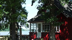 A red wooden cabin with a large window and a door, surrounded by trees and a body of water. - Charming Coastal Retreat in Zierow (Zierow)