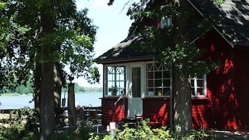 Eine rote Holzhütte mit einem großen Fenster und einer Tür, umgeben von Bäumen und einem Gewässer.