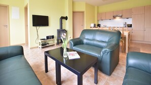 Een woonkamer met een bank, een salontafel en een televisie. Er is een keukenhoek met kastjes en een fornuis.