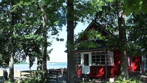 Een rood houten gebouw met veranda, omringd door bomen en uitzicht op het water.