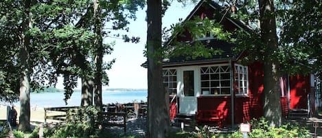 A red wooden building with a porch, surrounded by trees and a view of the water.