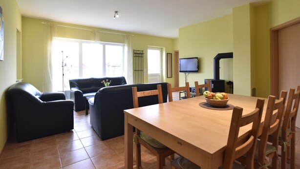 Een eetgedeelte met een houten tafel en stoelen, een zwarte bank en een televisie aan de muur.