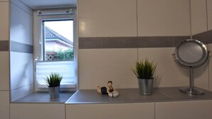 Ein Badezimmer mit Fenster, zwei Topfpflanzen, einer kleinen Figur und einem runden Spiegel an einer gefliesten Wand.