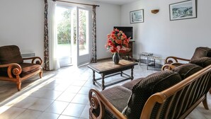 A living room with a sofa, armchairs, a coffee table, and a TV. There is a door leading to a garden.