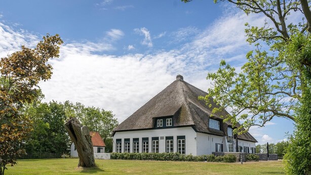 Een traditioneel rieten huis met witte muren en meerdere ramen, omgeven door een verzorgd gazon en bomen.