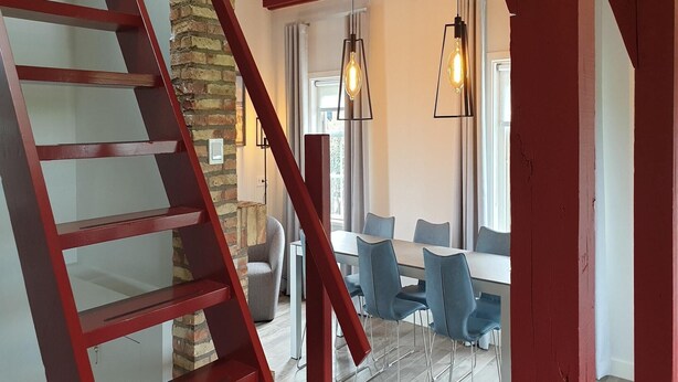 Un escalier avec des rampes rouges mène à un coin repas où se trouvent des chaises bleues et une table.