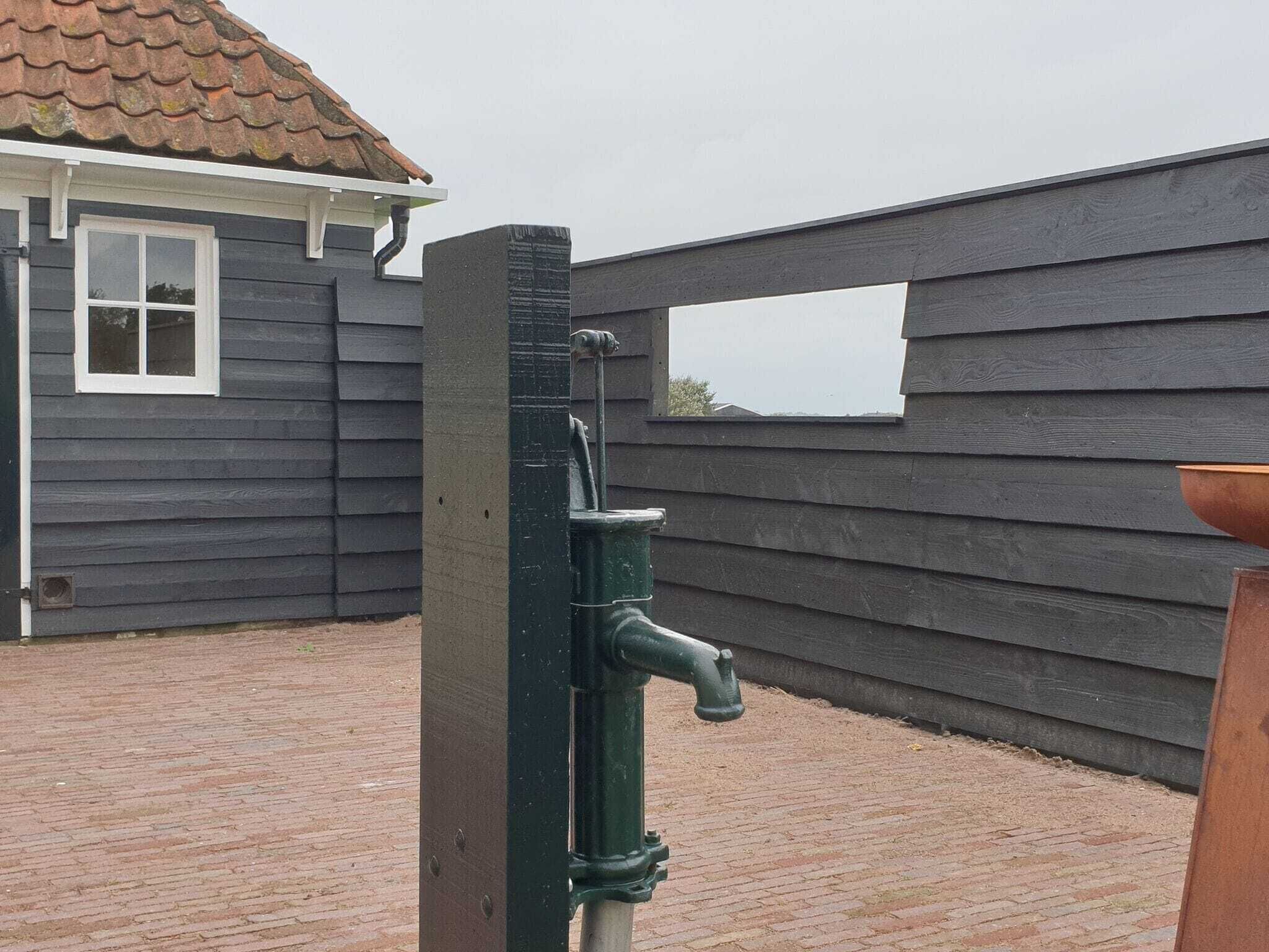 Un robinet en métal fixé à un poteau en bois, avec une terrasse en briques et une maison en arrière-plan.