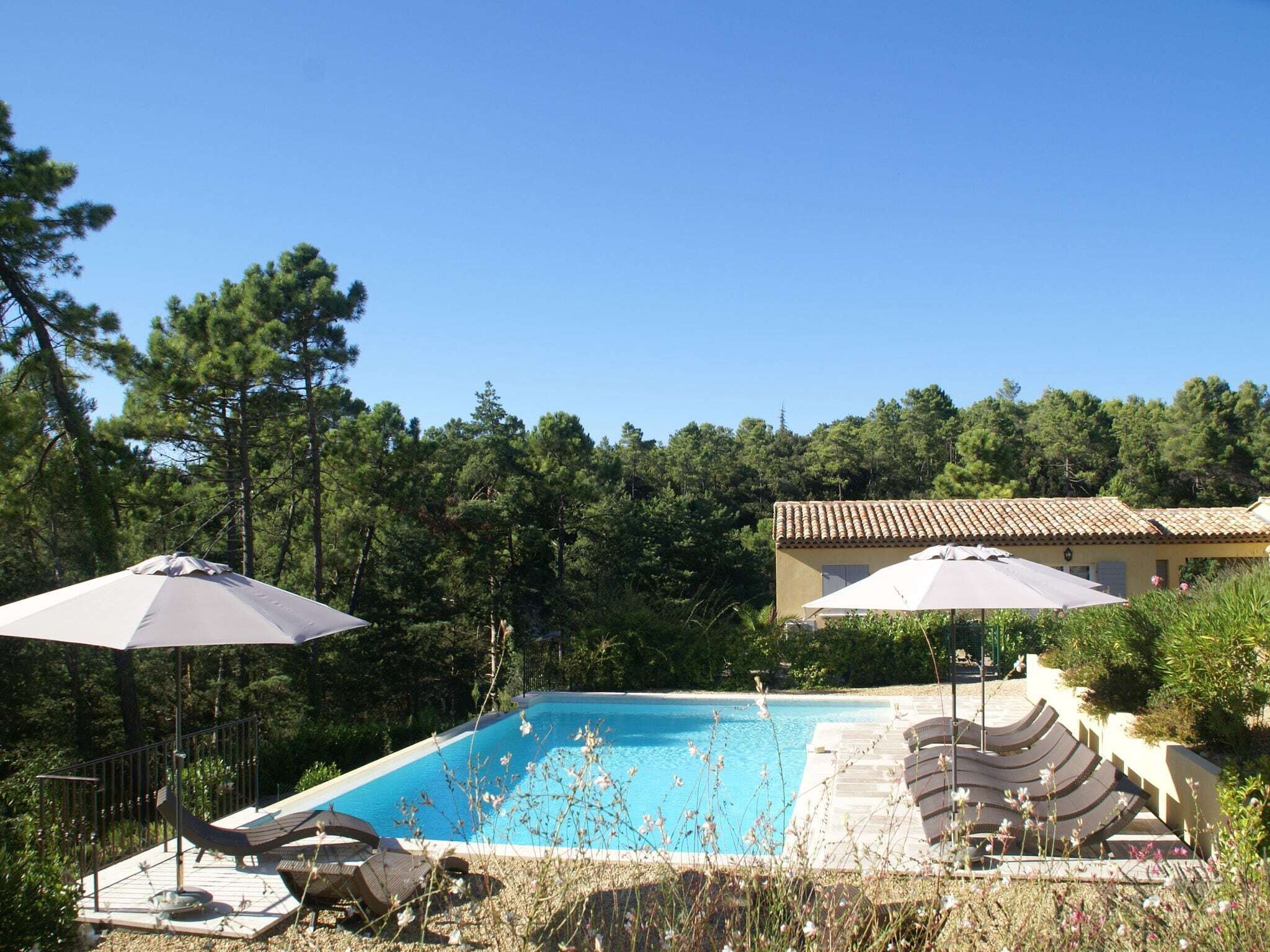 Una piscina con sillones de descanso y sombrillas, rodeada de árboles y un edificio con techo de tejas.