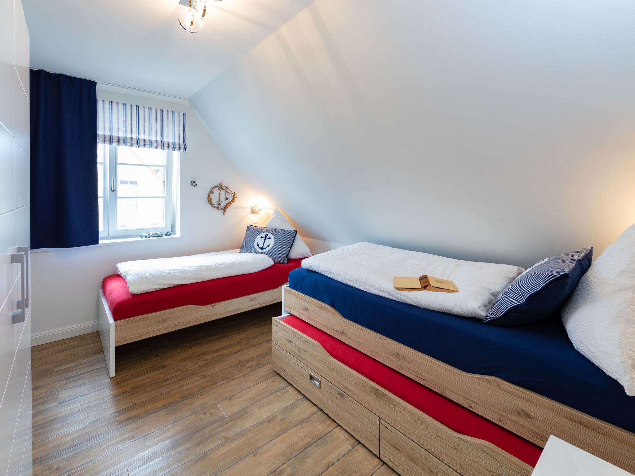 A bedroom with two beds, a window with curtains, a wall-mounted light, and a wooden floor.