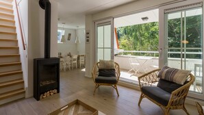 Ein geräumiges Wohnzimmer mit Kamin, Korbmöbeln und großen Schiebeglasfenstern, die auf einen Balkon mit Blick ins Grüne führen.
