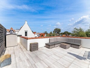 A rooftop terrace with a seating area, a wooden wall, and a view of neighboring buildings.