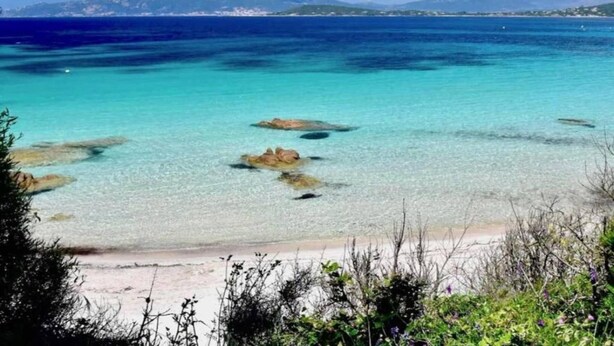 Ein Strand mit klarem türkisblauem Wasser, sichtbaren Felsen und einer fernen Küstenlinie mit Bergen.