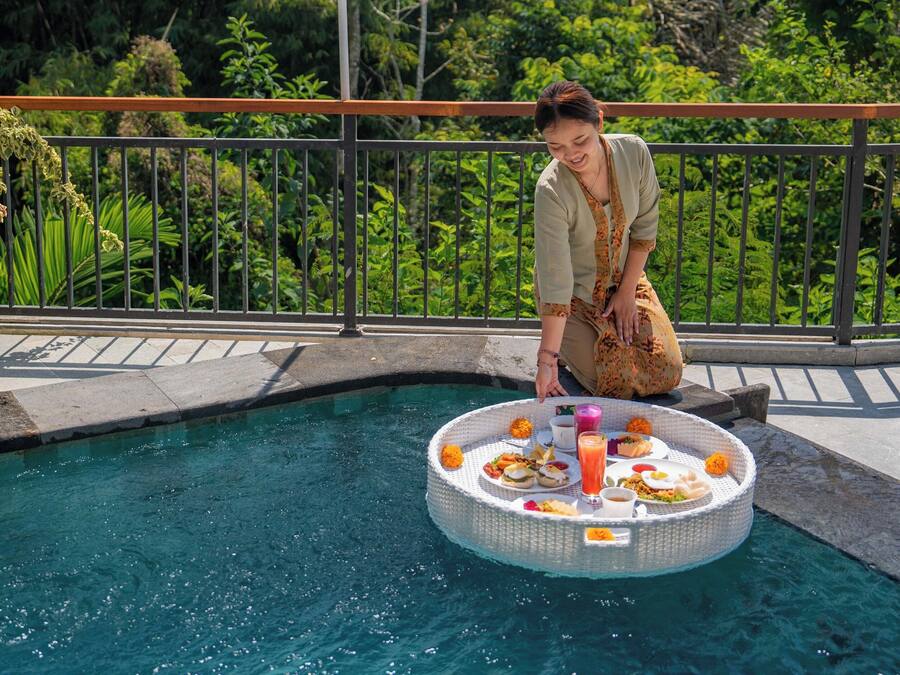 A poolside area with a floating table set with food and drinks, surrounded by a metal railing and lush greenery.