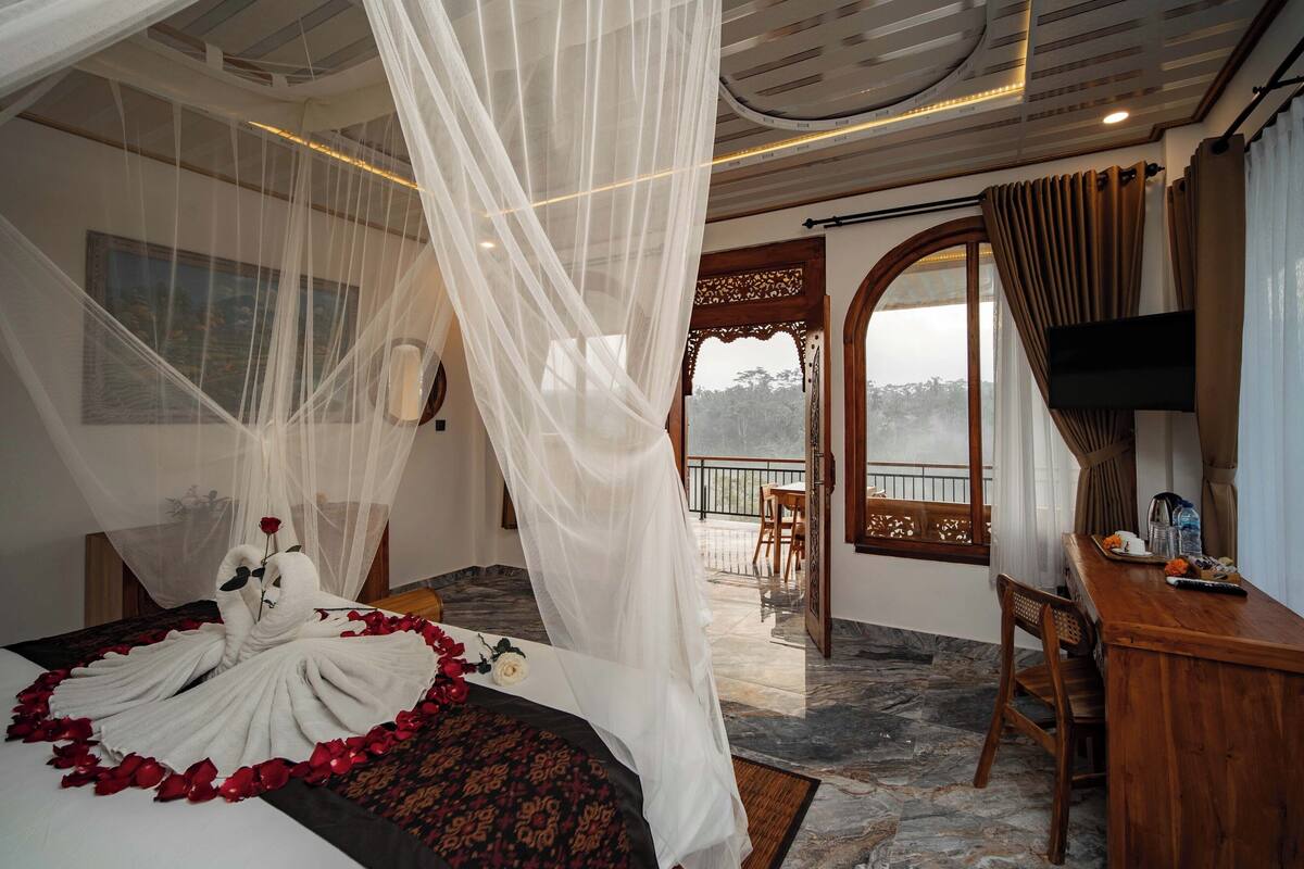 Quarto com cama decorada com pétalas de rosa e arranjo de toalha em forma de cisne, varanda com vista e escrivaninha de madeira com cadeira.