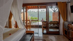 A bedroom with a bed, a bench, a small table, and a TV. There is a balcony with a view of greenery. - Campuhan Sebatu Resort (Sebatu)