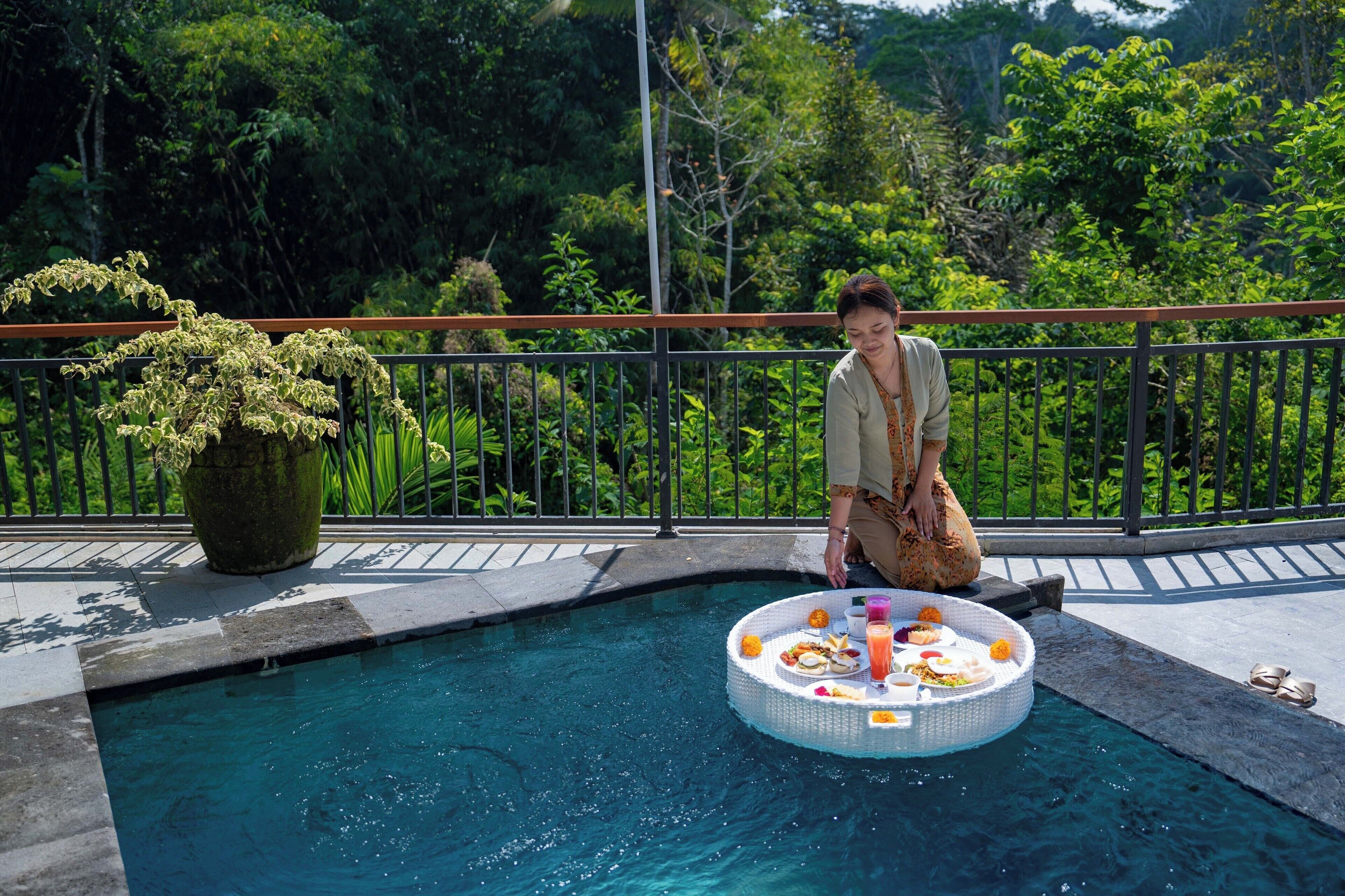 A poolside area with a circular floating table holding food and drinks, surrounded by lush greenery.