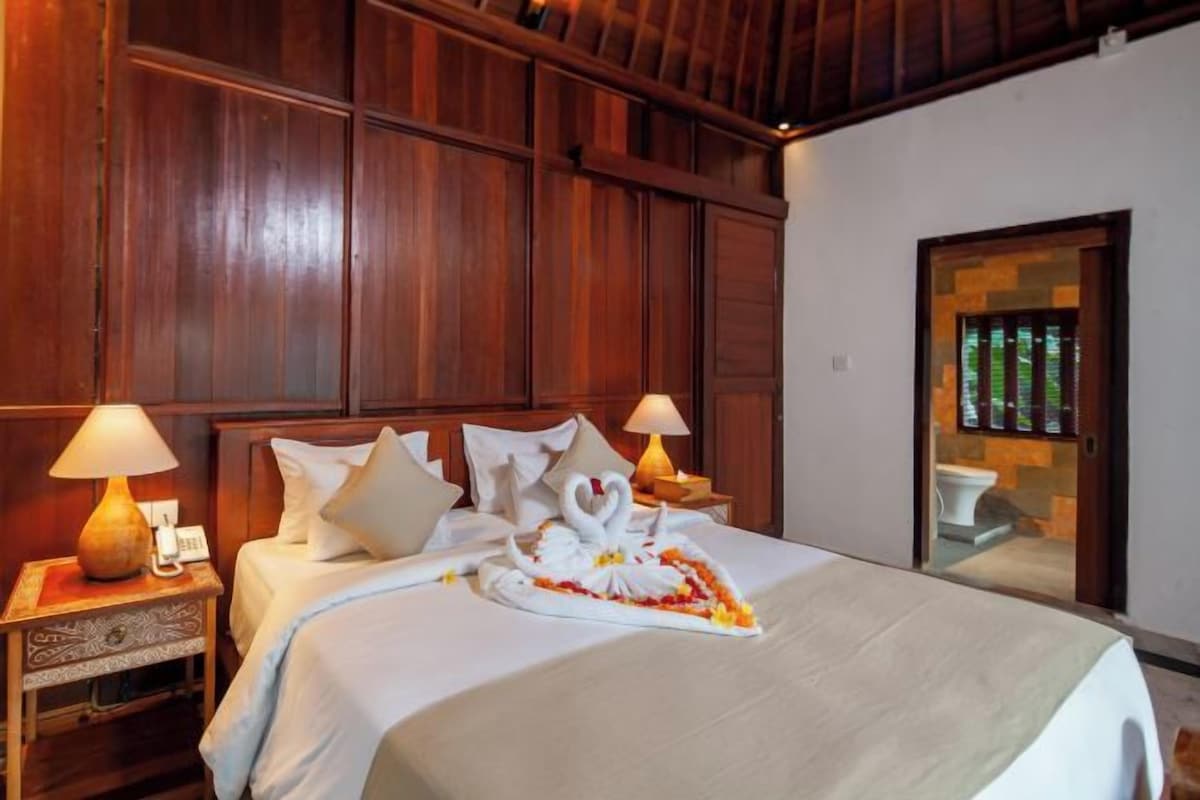 A hotel room with a wooden paneled wall, a bed with white bedding, two bedside lamps, and a swan-shaped centerpiece on the bed.