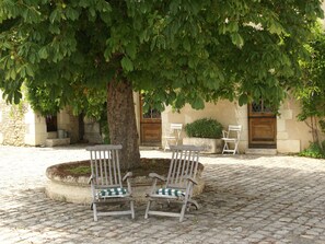 A courtyard with a large tree, two wooden chairs with cushions, and a stone wall.