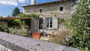 Una casa de piedra con chimenea, una ventana pequeña y una puerta con contraventanas. Hay sillas y una mesa al aire libre, bajo un toldo.