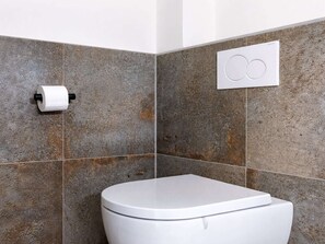 A bathroom with a white toilet, a toilet paper holder, and a wall-mounted flush button. - Apartment in Vrouwenpolder near the Beach (Vrouwenpolder)
