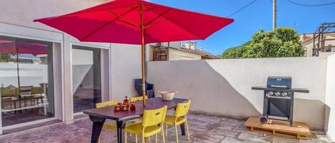 An outdoor patio with a table and chairs, a red umbrella, and a barbecue grill.
