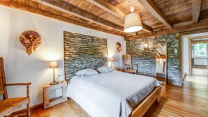 A bedroom with a stone wall headboard, wooden flooring, a large bed, a wooden chair, and a nightstand with a lamp. - Riverside Villa with Charm (Léglise)