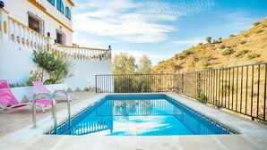 Una piscina con borde de baldosas, un edificio blanco con detalles azules y una colina al fondo.