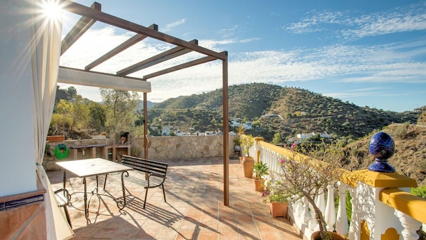 Un patio con mesa y banco, una pérgola, plantas en macetas y vistas a un paisaje montañoso.