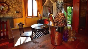 A rustic room with a stone wall, a wooden table with a bottle of champagne and a pineapple, and a view of greenery outside.