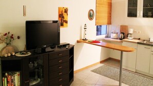 A compact living space with a TV on a dark wooden cabinet, a small kitchen area with a coffee maker, and a window with blinds. - Bungalow auf der Insel Poel for 2 Personen (Insel Poel)