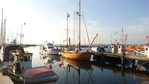 A marina with various boats docked, including a wooden boat with a tall mast and a white boat with a red cabin. - Bungalow auf der Insel Poel for 2 Personen (Insel Poel)