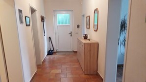A hallway with a tiled floor, a door at the end, and a wooden cabinet with a mirror and picture frames on the wall.