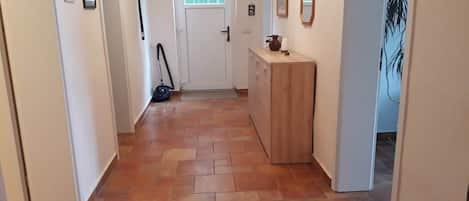 A hallway with a tiled floor, a door at the end, and a wooden cabinet with a mirror and picture frames on the wall.