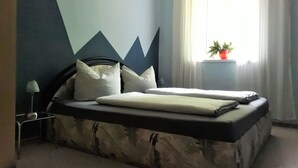 A bedroom with a bed, a nightstand, a lamp, a window with curtains, and a potted plant.