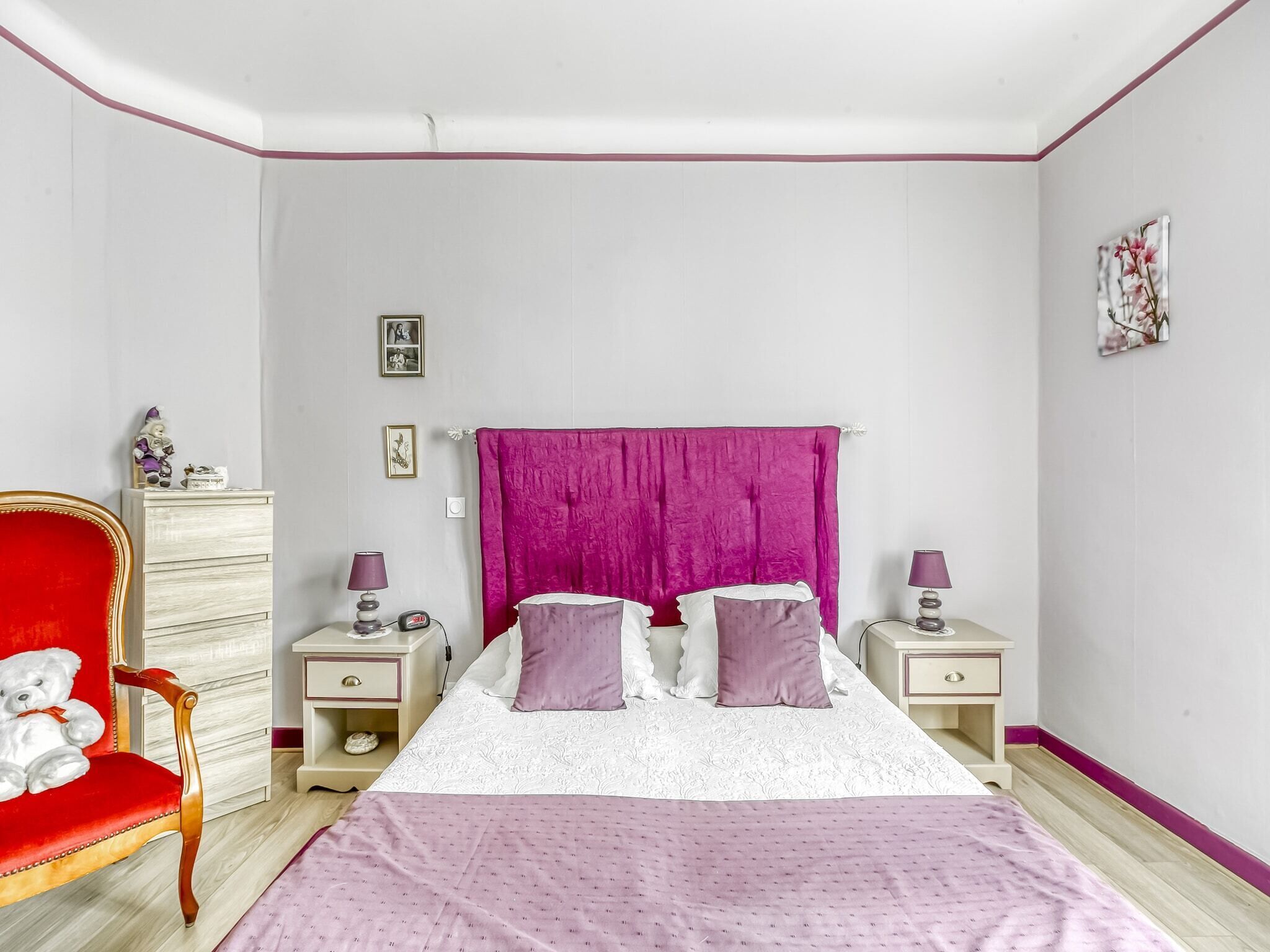 Un dormitorio con una cama, dos mesitas de noche, una silla roja, un aparador y una pared con un cuadro.