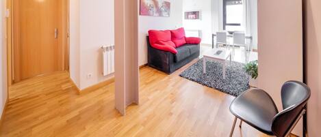 A modern living room with a wooden floor, a black sofa with red cushions, a black armchair, and a dining area with a table and chairs.