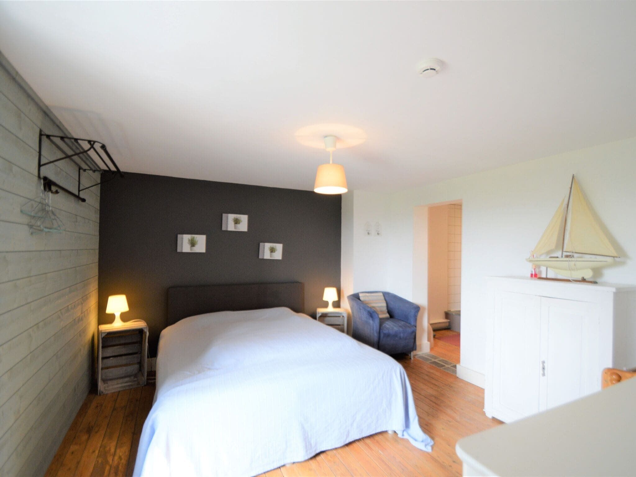 A bedroom with a bed, two bedside tables with lamps, a chair, a sailboat decoration, and a wardrobe.
