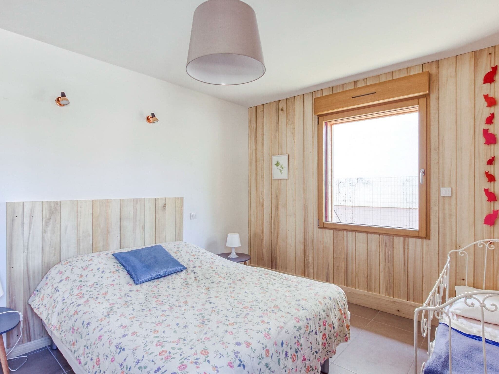 Un dormitorio con cabecera de madera, colcha estampada con motivos florales, almohada azul, mesita de noche con lámpara y ventana con vistas.