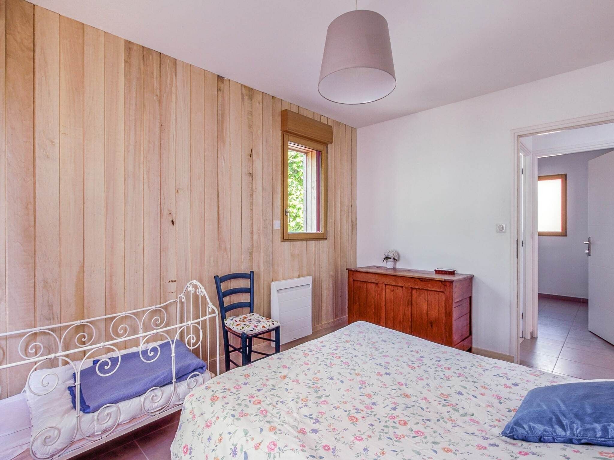 A bedroom with a wooden wall, a bed with a floral bedspread, a desk, a chair, and a hanging light fixture.
