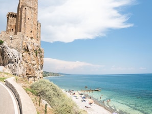 A coastal view with a historic stone tower and a beach with clear waters and umbrellas.