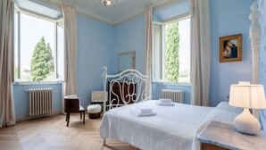 Un dormitorio con cama, mesita de noche, silla, cómoda, espejo y dos ventanas con cortinas.
